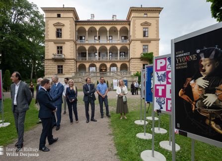 fotografia. art piknik w willi decjusza. ujęcie budynku willi od frontu. po dwóch stronach żwirowej alejki wystawa plakatów teatralnych, pomiędzy nimi spacerują goście.